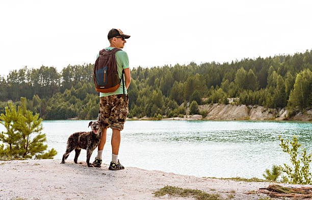 Caucasian man with a backpack and a Bedlington Terrier mix dog on white sand, looking at a turquoise lake. Concept of travel and adventure with a pet