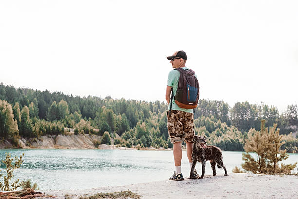 Man with backpack and mixed breed dog looking at a lake surrounded by forest. Summer travel and adventure concept for outdoors, walking and traveling with pet