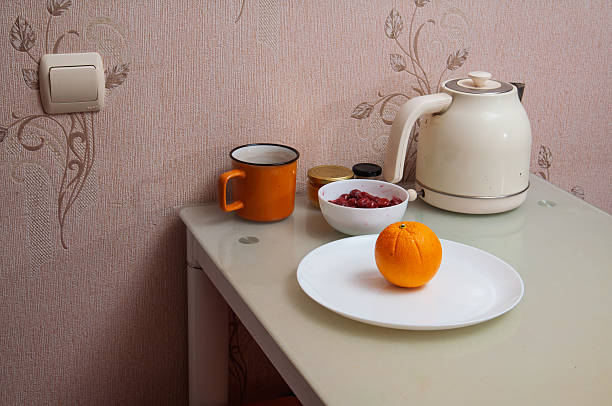 A still life scene featuring an orange on a plate, a bowl of berries, a mug, and an electric kettle on a glass table, with a light switch on the wall