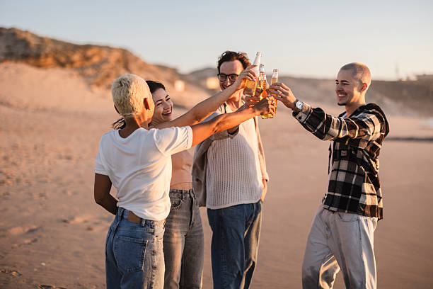 Group of diverse young friends cheers with bottles of refreshing drinks during golden hour on a sandy beach, embodying concepts of friendship, celebration, and leisure