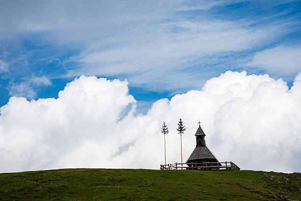 A traditional small wooden chapel with a cross sits on a green grassy ridge under a bright blue sky with large white fluffy clouds in Slovenia