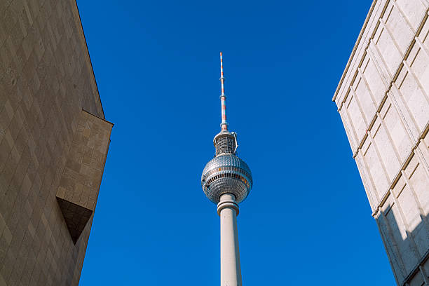 A striking view of Berlin's Fernsehturm rising between beige urban blocks against a vivid blue sky, highlighting modern architecture, cityscape, and towering height.