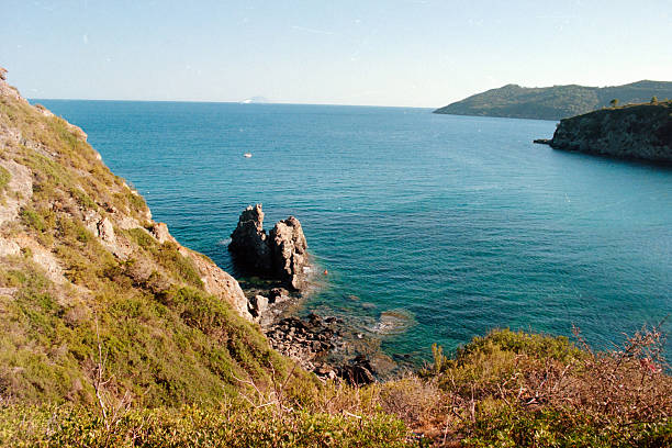 Azure Mediterranean Sea water washing against natural rock formations and green coastal hills under a clear blue sky. Scanned from analog film