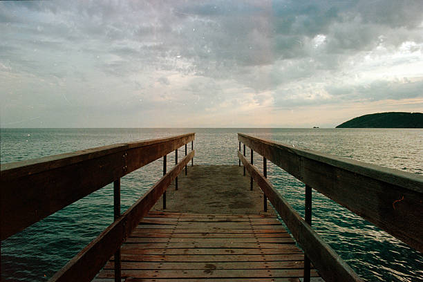 Wooden pier stretching towards the horizon, offering a peaceful view of the sea and distant island under a cloudy sky. Scanned from analog film