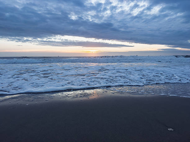Pre-dawn beach scene featuring a receding wave with the sun just below the horizon, casting a bluish glow. The soft foam of the wave stretches across dark, wet sand, reflecting the subtle colors of the sky. Overhead, scattered clouds add texture to the vast expanse, with a hint of light in the distance. The tranquil setting captures the peaceful transition from night to day at the coastline.