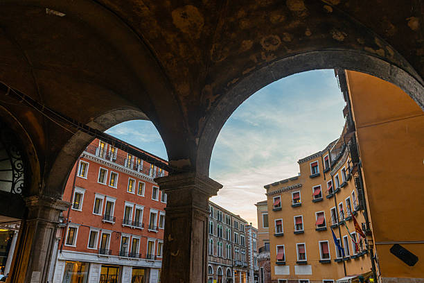 View of historic buildings in Venice, Italy, framed by stone arcade arches. Colorful facades, traditional windows and architectural details illustrate the characteristic urban landscape of the Venetian historic center under soft evening light.