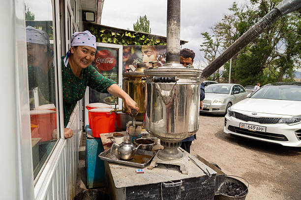 Smiling woman serves drinks from large metal samovar at roadside stall. Busy atmosphere with cars and cheerful mood in daytime setting in Cholok, Kyrgyzstan - July 16, 2024