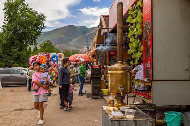 Five people, including a woman carrying a baby, engage in lively activities at an outdoor roadside cafe. Colorful stalls filled with food, drinks, and festive items create a vibrant atmosphere under a partly cloudy sky in Cholok, Kyrgyzstan - July 16, 2024