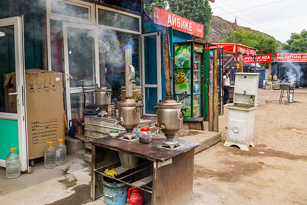 Woman looks away while samovars emit steam at bustling roadside roadside cafe. Dusty ground and shops create a warm, rustic atmosphere in Cholok, Kyrgyzstan - July 16, 2024