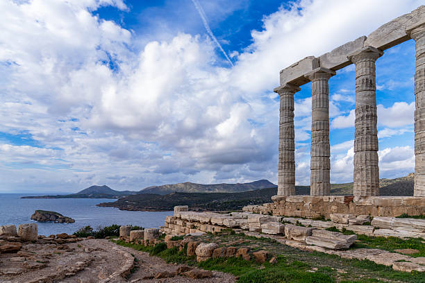 A horizontal wide-angle perspective showing the weathered marble columns of the ancient Temple of Poseidon against a dramatic cloudy sky, with the coastline and a small island in the background.