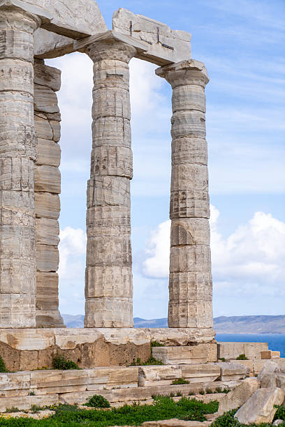 A vertical medium shot focusing on the weathered texture and impressive scale of the Doric columns at the Temple of Poseidon, Cape Sounion, set against a bright sky.