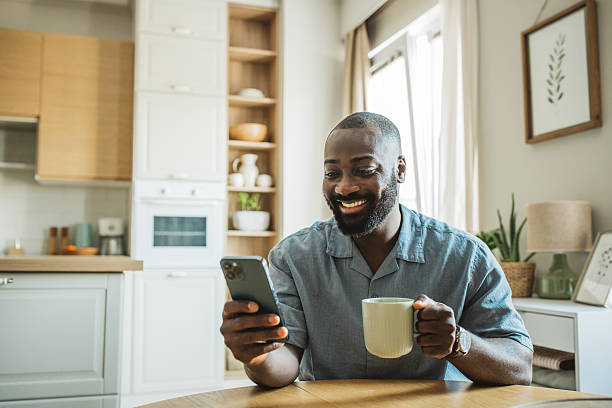 istock Carefree black man using smartphone and drinking coffee while sitting at kitchen 2260117893