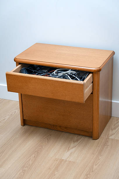 Open wooden drawer filled with a disorganized tangle of electronic cables against a white wall