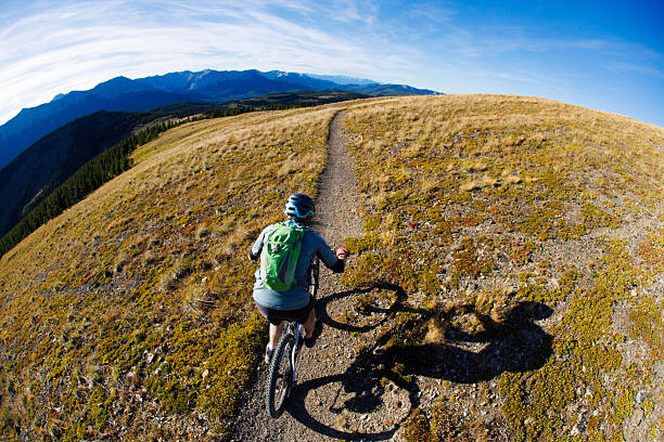 A man rides his electric-powered mountain bike down a singletrack trail in the Rocky Mountains of Alberta, Canada, on a sunny afternoon. He wears cycling clothing, a backpack, and a bicycle helmet.