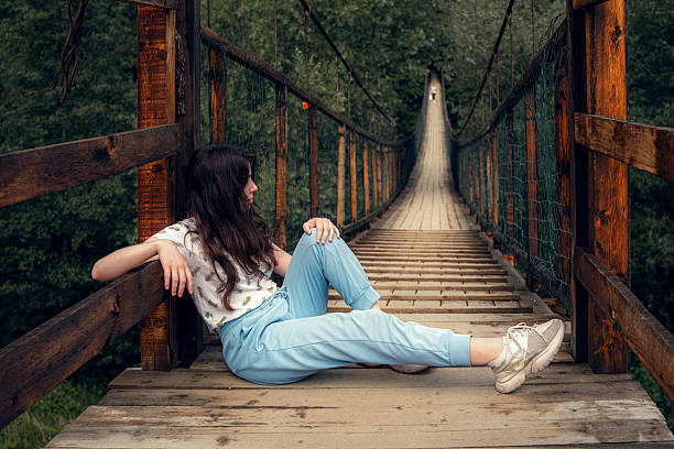 A young woman sitting and posing on a wooden suspension bridge surrounded by green forest. She is looking at the camera with a calm and thoughtful expression. The image represents solitude, travel, freedom, and connection with nature.