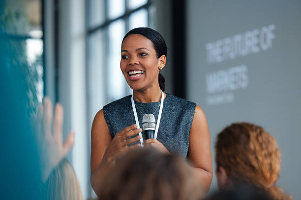 A confident female speaker presents an engaging talk at a conference, addressing a crowd of attendees. The scene emphasizes business, education, professional communication, and audience engagement in a modern event setting.