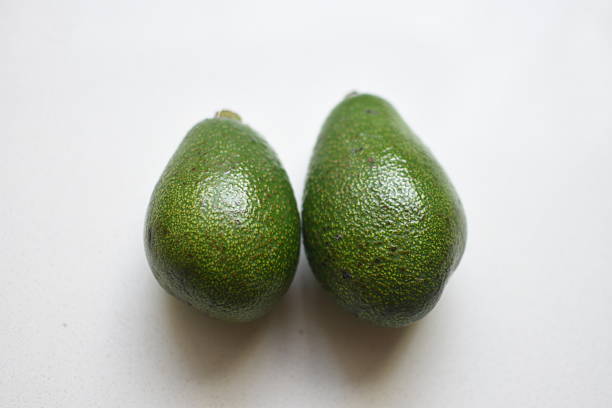 Two fresh avocados placed on a white table, highlighting their vibrant green color and natural texture for culinary, food photography, and healthy lifestyle concepts.