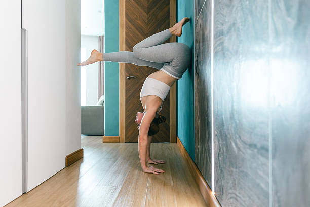 Athletic woman performing advanced wall-supported handstand yoga indoors. Concept of strength, balance, flexibility, body control, home workout, mindful fitness and healthy lifestyle.