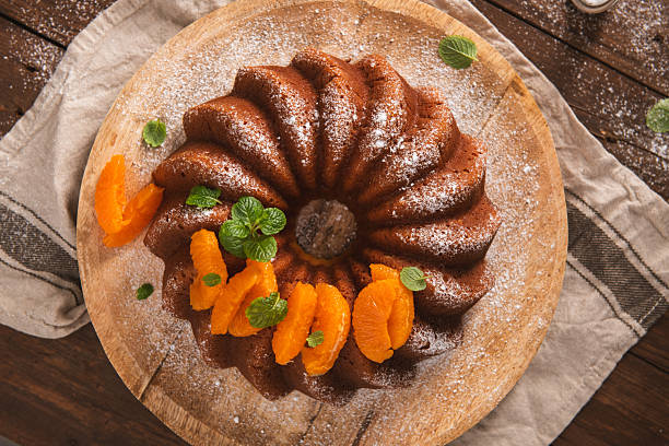 A top-down view of a golden-brown citrus bundt cake dusted with icing sugar. Decorated with vibrant orange segments and mint leaves, the scene features warm lighting and rustic textures.