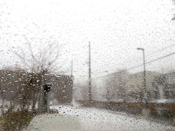 Raindrops covering a window on a blurry, overcast day looking out onto a snowy urban street