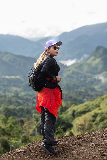 A woman is hiking, standing on a cliff with forest and mountains in the background.