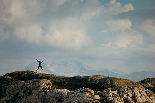 Distant view of woman trail runner jumping for joy on mountain ridge, lofty clouds and snowcapped mountain behind, Whistler, B.C.