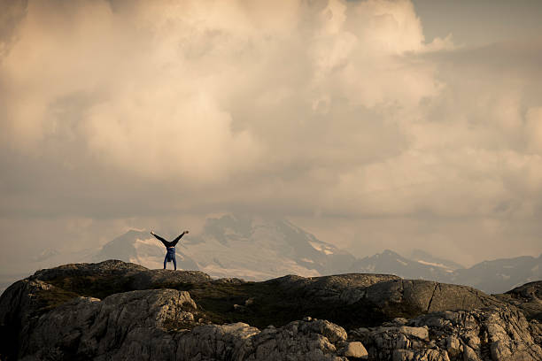 Distant view of woman trail runner performing handstand on mountain ridge, lofty clouds and snowcapped mountain behind, Whistler, B.C.
