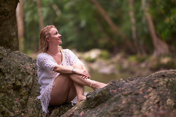 Happy mid adult woman day dreaming while relaxing on a rock during summertime in the forest.
