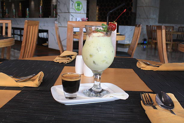 Refreshing green smoothie garnished with a cherry and a shot of espresso on a dining table in a restaurant Cutlery wrapped in yellow napkins completes the place setting