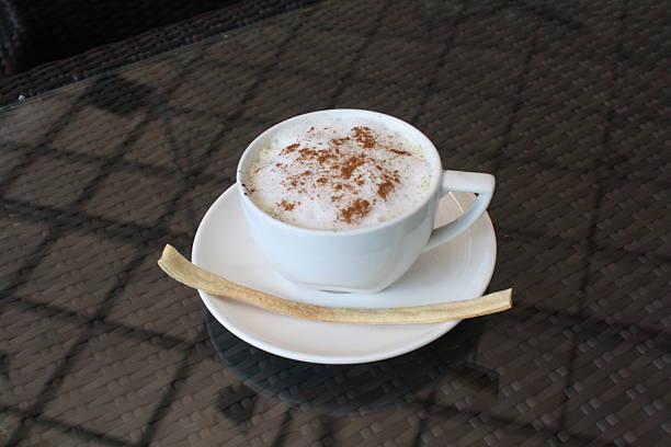 Frothy cappuccino with a sprinkle of cinnamon, served in a white cup with a crispy biscuit on a glass topped weave table