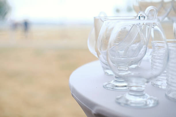 Clear glass cups are arranged on white tablecloth with blurred outdoor background showing people walking and grass area in distance with soft lighting