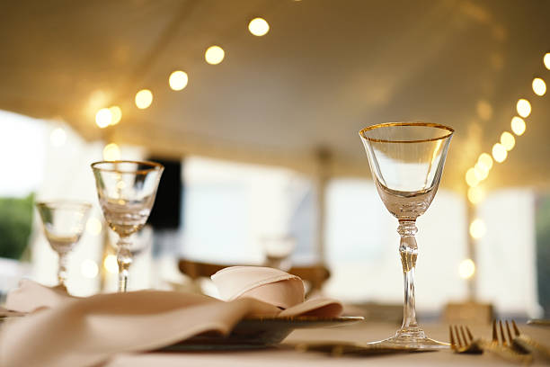 Glassware arranged on table with white napkin and silver cutlery under tent with string lights in background creating soft lighting atmosphere
