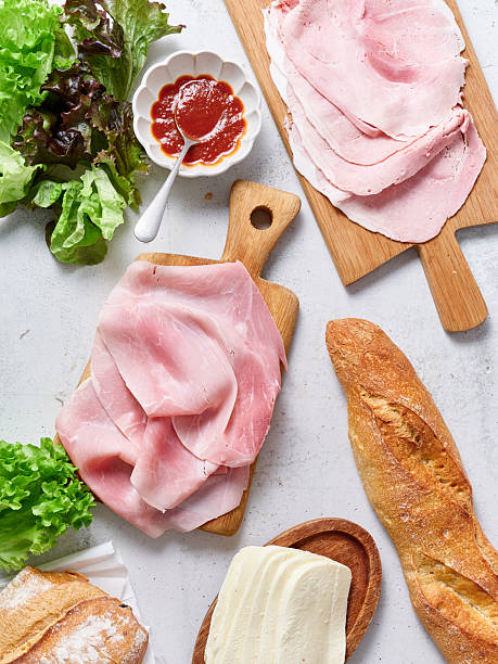 Lunch setup with fresh ingredients for sandwiches including ham, mozzarella, lettuce, and baguette by a table with oranges and scrambled eggs