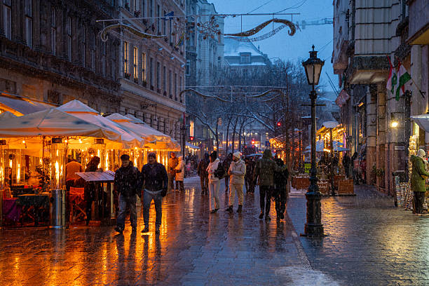 Budapest, Hungary - January 6, 2026: Pedestrians stroll through a snowy Budapest street at night, where cozy outdoor cafes with warm lights and heaters contrast against the blue twilight and falling snow.