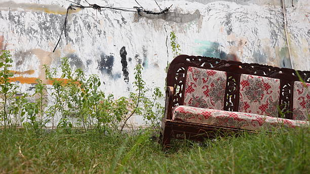 A forgotten wooden couch with red patterned upholstery lies abandoned on grass beside a peeling graffiti wall, evoking stories of loss, decay, and neglect.