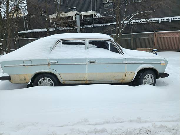Kyiv, Ukraine - February 5 2026: A vintage Soviet light-blue sedan car sits buried in deep snow. Heavy white drifts cover its roof and hood, while the lower body shows significant rust. It’s parked in a wintry urban setting with bare trees and a dark building in the background. A cold, quiet scene of seasonal neglect.