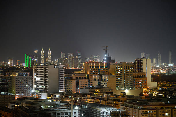 Urban residential blocks in the foreground contrast with Dubai's illuminated skyline at night. The city lights reflect off the modern skyscrapers, creating a vibrant urban landscape. Clear skies enhance the clarity of the buildings' outlines, while the overall scene depicts a bustling metropolis.
