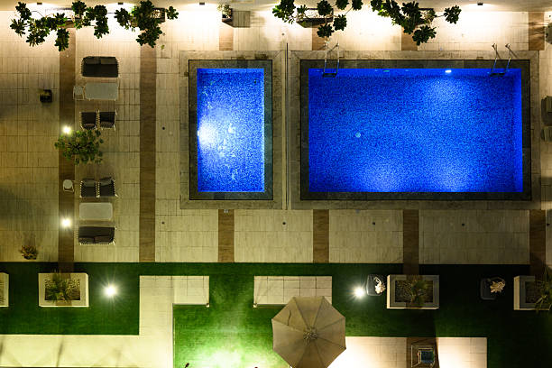 High-angle perspective looking directly down upon a luxury hotel rooftop area in Dubai. Two rectangular swimming pools glow intense electric blue, surrounded by light tiled decking and artificial green turf. Modern outdoor seating areas sit vacant under bright spotlights, creating a striking geometric pattern. The scene conveys upscale leisure and nocturnal hospitality.