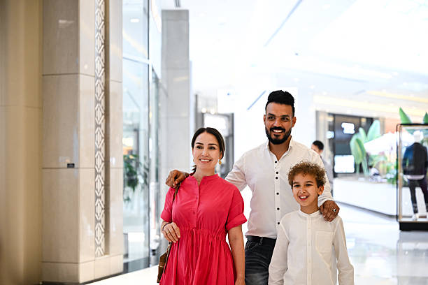 Middle-Eastern parents and young son walking side-by-side through a luxurious, bright shopping mall in Dubai. The father places his arm around the boy's shoulder, smiling warmly at the camera. Natural daylight streams through large glass windows, illuminating the polished stone floors and modern architectural details. The happy family enjoys quality time shopping.