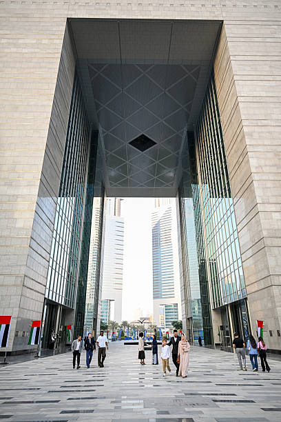 Groups of people walking through the massive shaded archway of The Gate Building at the Dubai International Financial Centre (DIFC). Modern stone and glass architecture creating a striking contrast against the bright midday sunlight illuminating the pedestrian plaza. Tall skyscrapers reflecting the clear sky defining this global business environment.