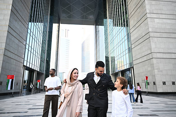 A family walking together through the DIFC Gate Building in Dubai, surrounded by modern architecture. The father, mother, and child engage in conversation as they enjoy their surroundings. The scene is set on a clear day with bright lighting, highlighting the impressive glass and stone structures.