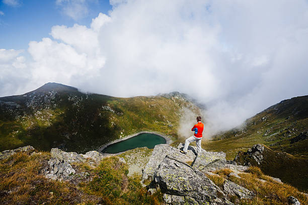 Young adult man stands on a rocky cliff above a small mountain lake, surrounded by steep slopes and moving clouds. Outdoor hiking scene with grass, stones, and open sky, captured in natural daylight.