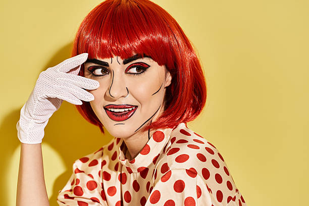 A pretty redhead woman in a polka dot shirt and white gloves, with creative pop art makeup, stands against a yellow background.