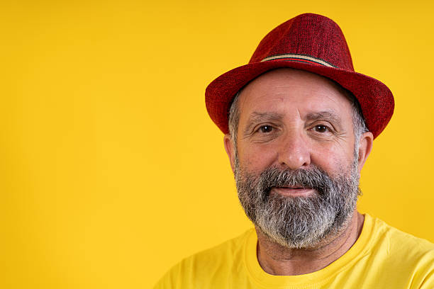 Mature bearded man in a red fedora smiling confidently in a bright yellow studio portrait, head-and-shoulders, looking at camera