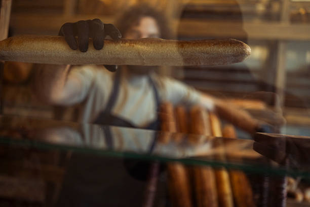 Male baker carefully arranges baguette loaves in the bakery display window, presenting freshly baked bread to customers
