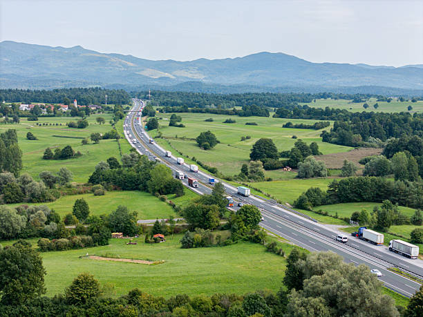 Aerial view of a highway with trucks and cars winding through green farmland and trees, with distant mountains under a cloudy sky.