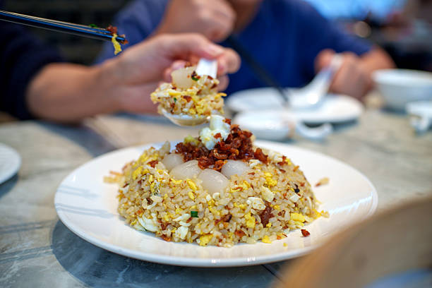 Close-up of a person using chopsticks to eat fried rice topped with scallop, served on a white plate at a casual restaurant. Asian dining culture, everyday eating moments, and freshly prepared comfort food in a social setting.