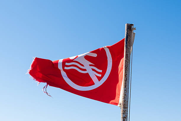 Red flag featuring a white emblem waves in the wind, mounted on a weathered pole against a bright blue sky backdrop