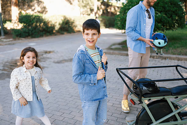 Mother enjoying a leisurely stroll in a paved urban park, pushing a stroller with her toddler, while her older son cycling alongside, creates a scene of family bonding and happiness