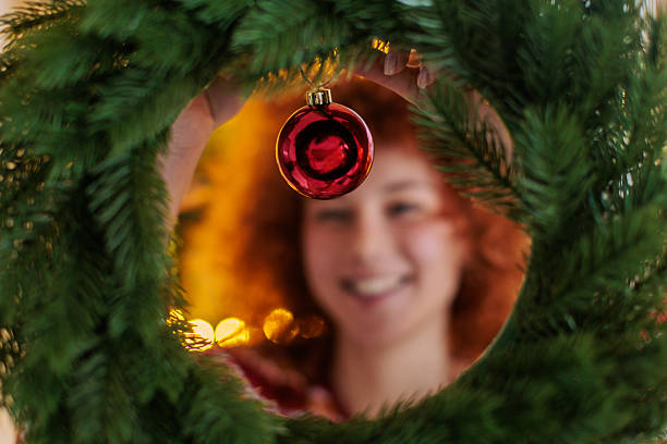 A smiling woman decorates a Christmas wreath indoors, holding festive ornaments while looking through the wreath. The close-up portrait captures a cozy holiday atmosphere with warm lights, seasonal decorations, and a cheerful Christmas mood.